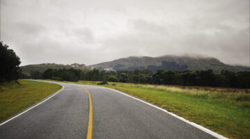 Under a heavy gray sky, a quiet road curves gently into the distance, leading toward misty hills cloaked in low clouds. The moody atmosphere, softened by the muted greens of the grass and trees, invites contemplation. On the horizon, a cluster of buildings nestles into the landscape, dwarfed by the looming, fog-shrouded mountains behind them. It’s a scene that whispers of slow journeys, hidden destinations, and the peaceful solitude of a road less traveled.